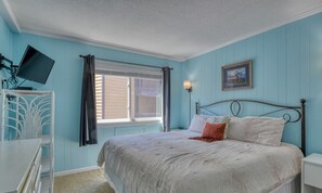 A bedroom with a bed, a desk, a chair, a window with curtains, and a wall-mounted TV.