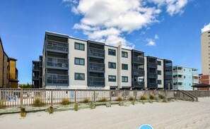 A multi-story residential building with balconies, a wooden fence, and a sandy beach in the foreground.