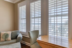 A room with a sofa, a patterned armchair, a wooden table, and large windows with blinds.