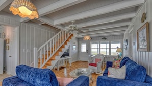 A spacious living room with a wooden staircase, blue upholstered furniture, and a large window with a view. - Love's A Beach - 5 Bedroom. 4 Bathroom, Oceanfront Dog Friendly Home (Garden City Beach)