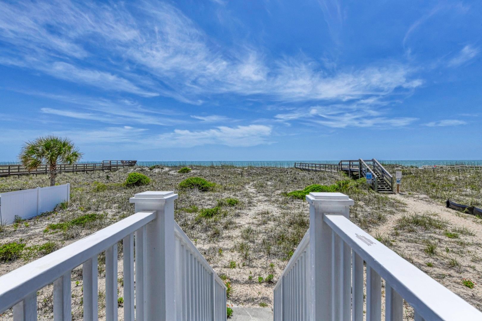 A beach pathway leading to the ocean with a wooden fence, a palm tree, and a wooden staircase.