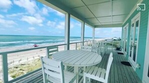 Ein überdachter Außenbereich mit Tisch und Stühlen, der einen Strand mit einem Zaun und eine Person in der Ferne blickt.