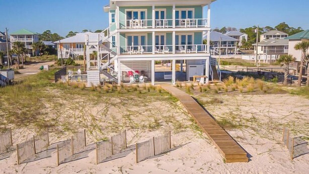 Una casa de playa de dos pisos con un porche cubierto y una terraza de madera que conduce a la entrada.