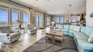 Un salón amplio con un gran sofá en forma de L, dos sillones y una mesa de centro. La habitación tiene ventanas grandes que ofrecen vistas al océano.