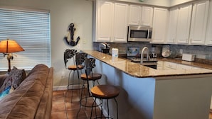 Una cocina con armarios blancos, encimera de granito y microondas integrado. Hay una zona con taburetes de bar, tres taburetes y una pequeña isla con un fregadero.