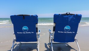 Two beach chairs with the logo of Garden City Realty, Inc. on the backrests, placed on a sandy beach with the ocean in the background.