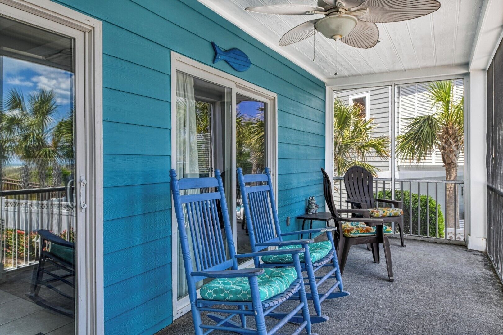 A covered porch with blue chairs and a table, a ceiling fan, and a view of palm trees.