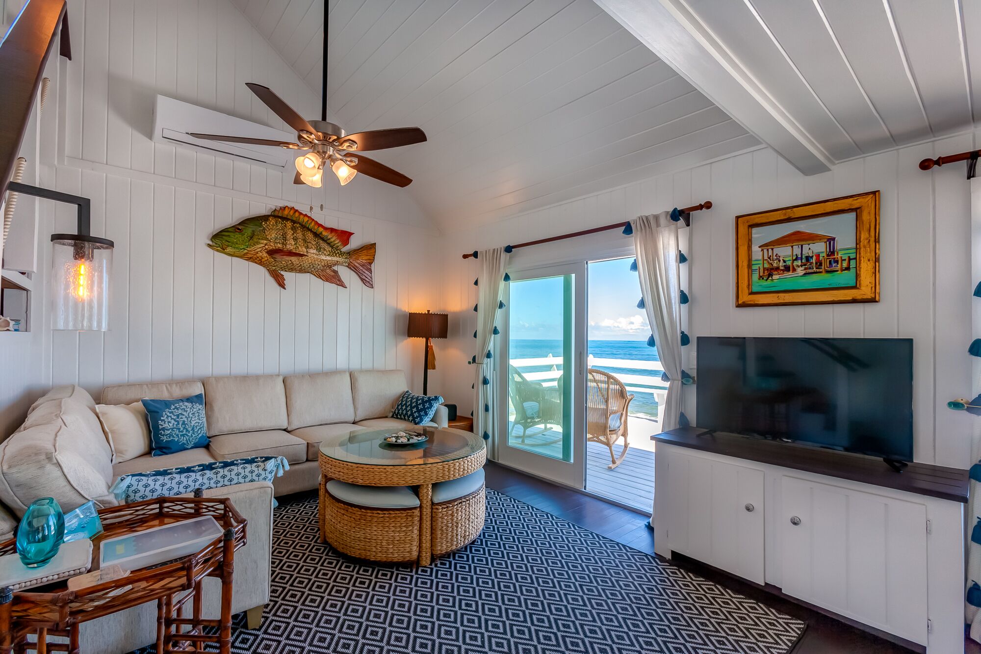 A cozy living room with a sofa, a round wicker table, a ceiling fan, and a large TV.