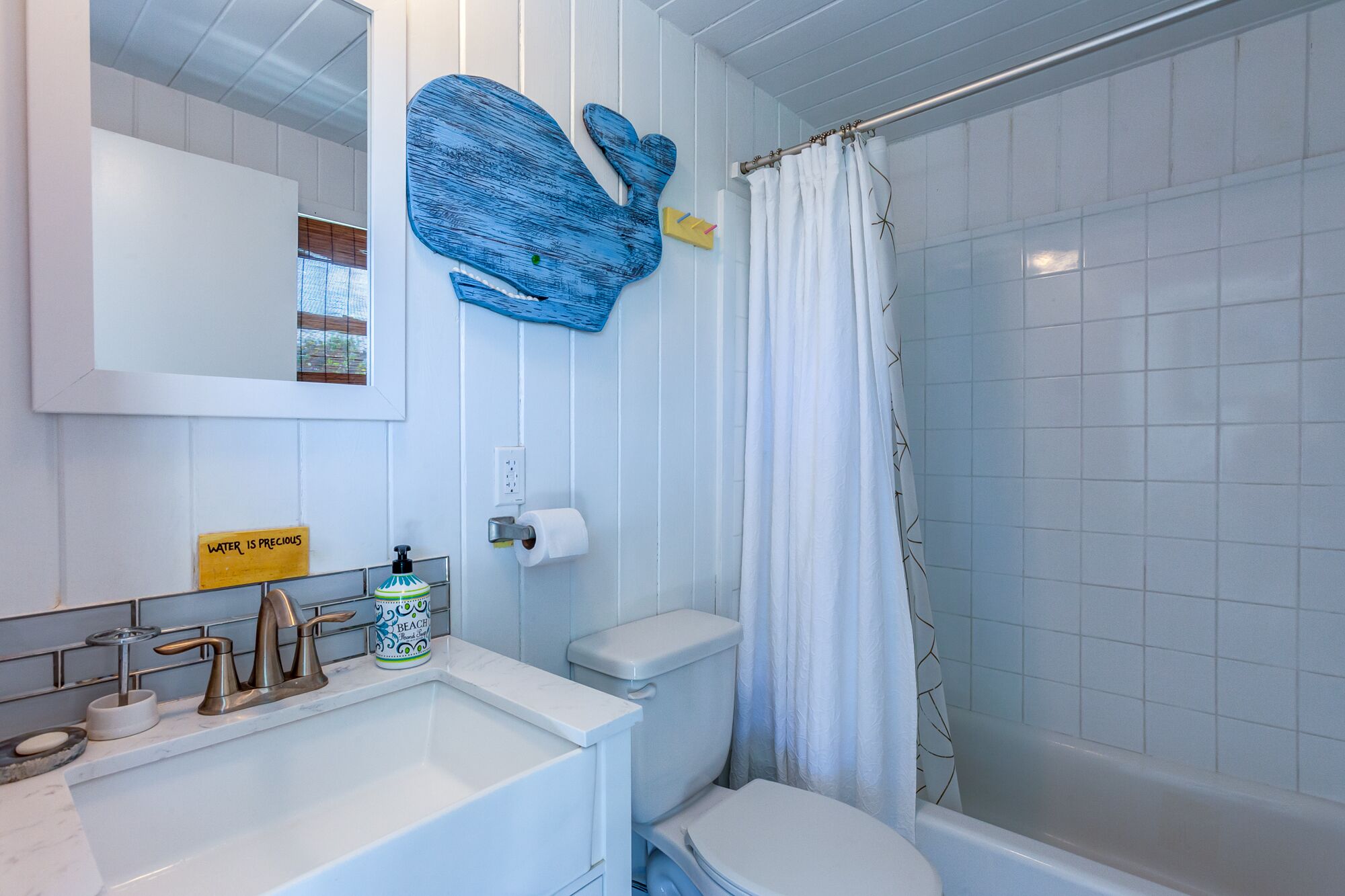 Une salle de bain avec un lavabo, des toilettes et une douche, le tout de couleur blanche. Un élément décoratif bleu orne le mur, et un panneau jaune indique : « L’EAU EST UNE RESSOURCE PRÉCIEUSE ».