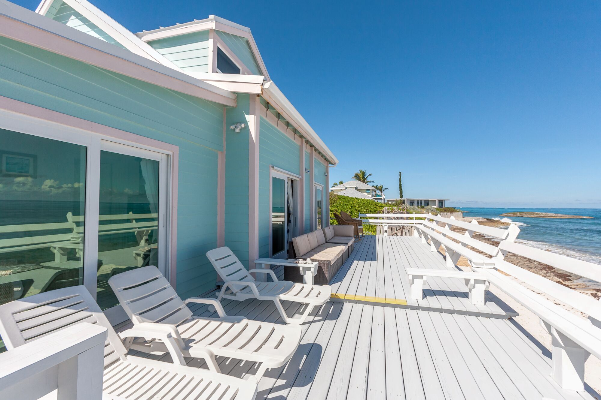 A beachfront property with a wooden deck, white lounge chairs, and a glass door leading to a covered area.