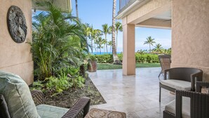 A covered patio area with wicker furniture, a decorative wall piece, and a view of palm trees and the ocean.