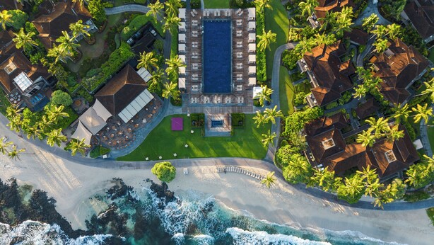 An aerial view of a resort with a central pool area, multiple guest villas, and a beachfront.