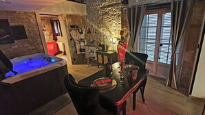 A cozy room with a stone wall, a wooden table with a red chair, a small table with a candle, and a window with shutters.