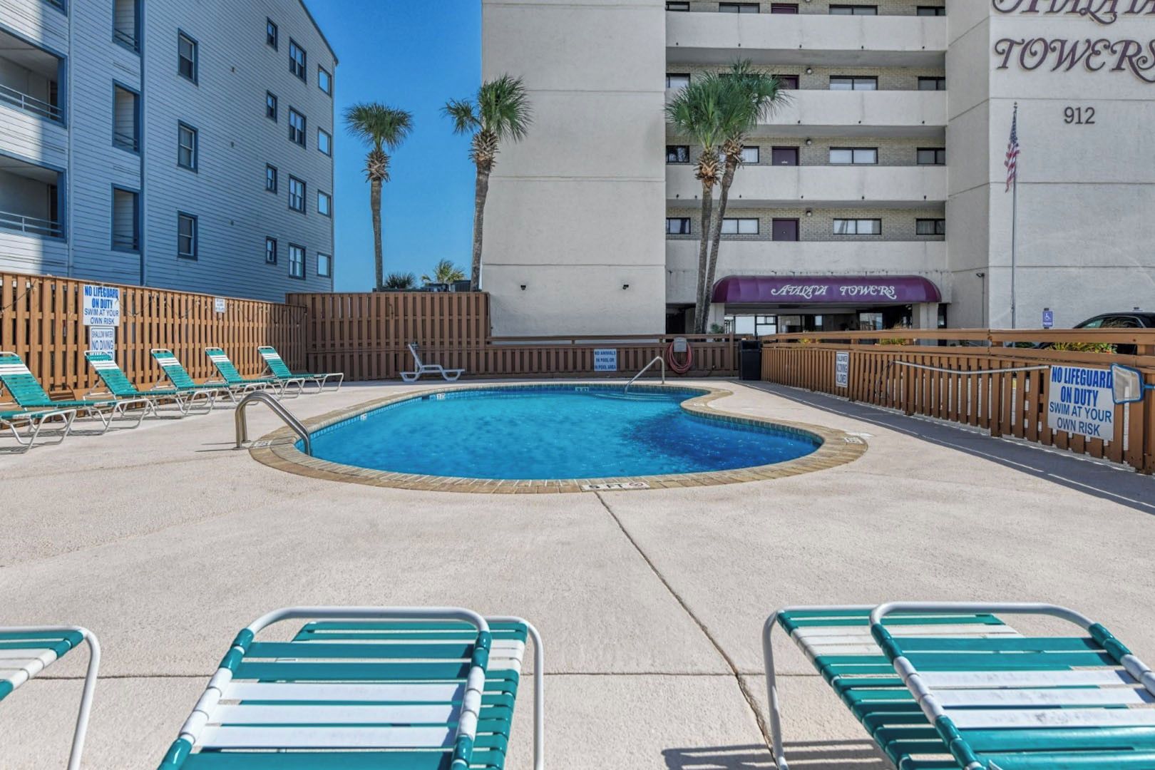 A swimming pool with lounge chairs and a wooden fence, surrounded by buildings.