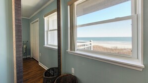Um corredor com um espelho grande refletindo a vista da praia, uma janela com vista para a praia e uma cesta de vime no chão.