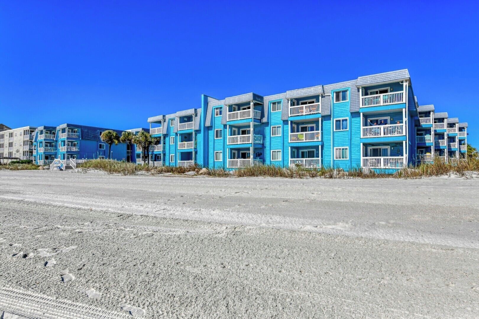 A beachfront hotel with multiple blue and white buildings, featuring balconies and a sandy beach in the foreground.