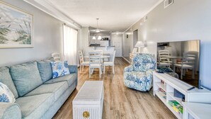 A modern living room with a grey sofa, a patterned armchair, a dining table with chairs, a TV, and a bookshelf. - Beach House 105 - Two Bedroom, Two Bath Updated Beach Condo (Garden City Beach)
