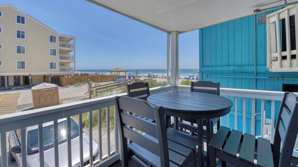 A balcony with a table and chairs, overlooking a beach with a wooden fence and a building in the background. - Beach House 110, two-bedroom, two-bath ocean-view condo (Garden City Beach)