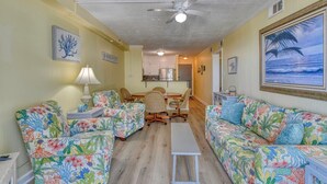 A living room with two floral-patterned sofas, a dining table with chairs, a ceiling fan, and a kitchen in the background. - Beach House 110, two-bedroom, two-bath ocean-view condo (Garden City Beach)