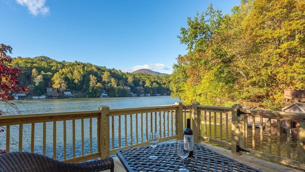 Une terrasse avec une table et des chaises donnant sur un lac, agrémentée d’une rambarde en bois et d’une bouteille de vin.