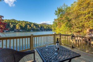 A deck with a table and chairs overlooking a lake, with a wooden railing and a bottle of wine. - Lake Lured Away | Dock, Game Room, Grill + Views! (Lake Lure)