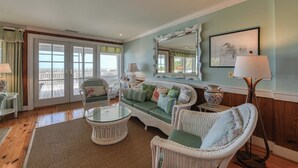 Un salon avec des meubles en osier, une table basse en verre, un canapé avec des coussins, un lampadaire, un miroir et un tableau encadré accroché au mur.