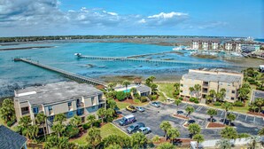 Une vue aérienne d’une zone côtière comprenant un port de plaisance, une longue jetée et plusieurs bâtiments, dont un hôtel avec un parking et une piscine.