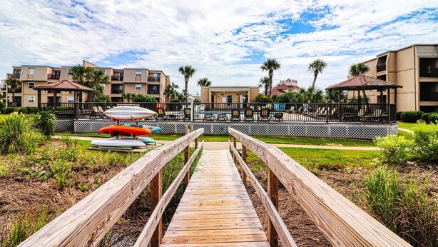 A wooden boardwalk leading to a resort area with outdoor seating and palm trees. - Inlet Pointe 33, two-bedroom, two-bath Inlet/channel-front condo (Garden City Beach)