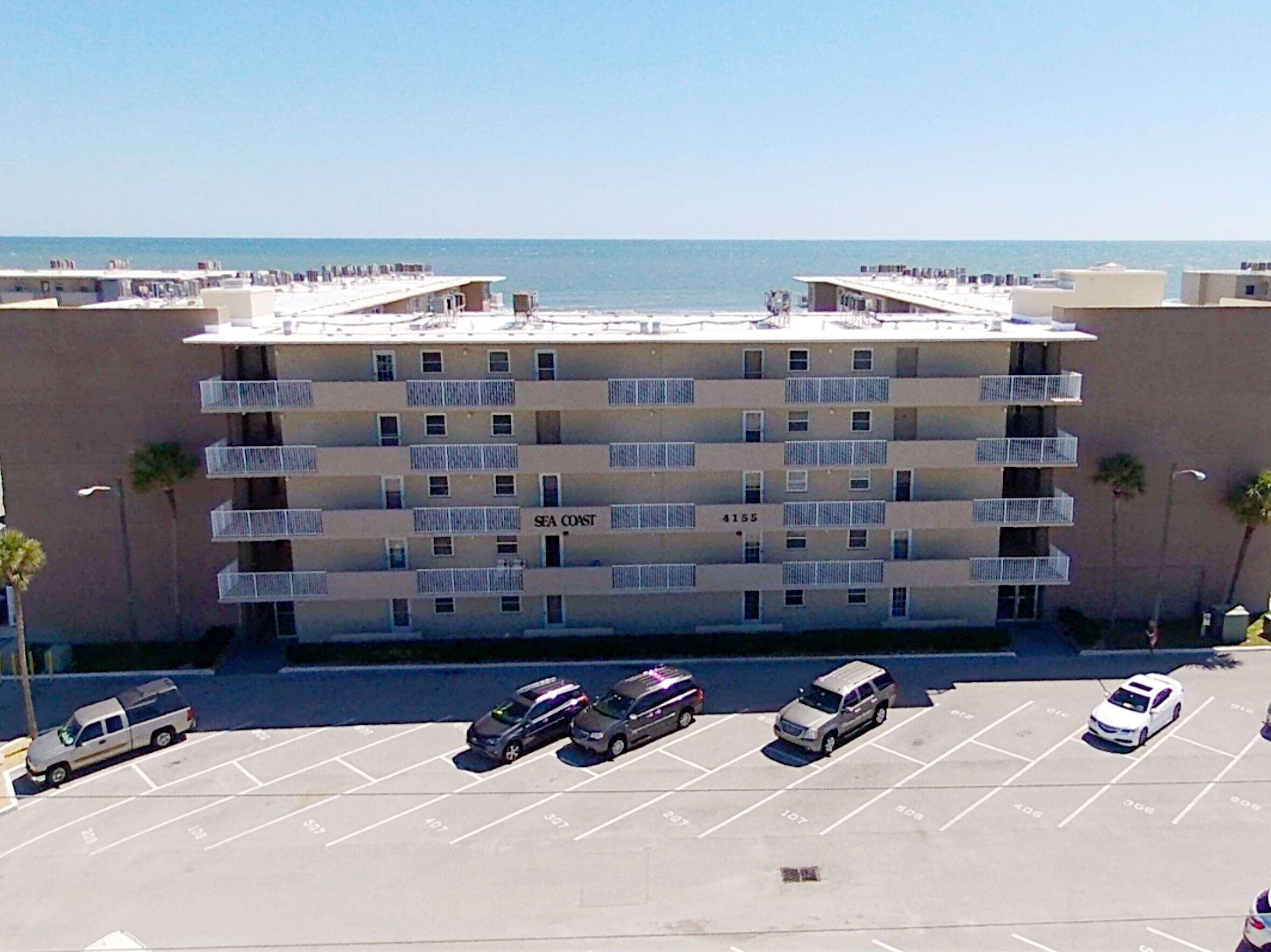 A multi-story hotel with balconies, a parking lot, and a view of the sea.