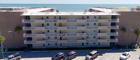 A multi-story hotel with balconies, a parking lot, and a view of the sea.