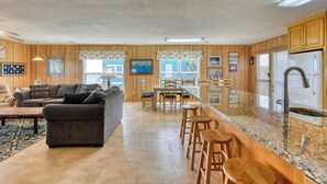 Un grand espace de vie comprenant un canapé, une table basse et une table à manger avec des chaises. Il y a également un coin cuisine équipé d’un réfrigérateur et de tabourets de bar.