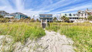 Une propriété située en bord de mer, avec un sentier sablonneux menant à un escalier en bois, entourée de hautes herbes et de maisons dotées de balcons.