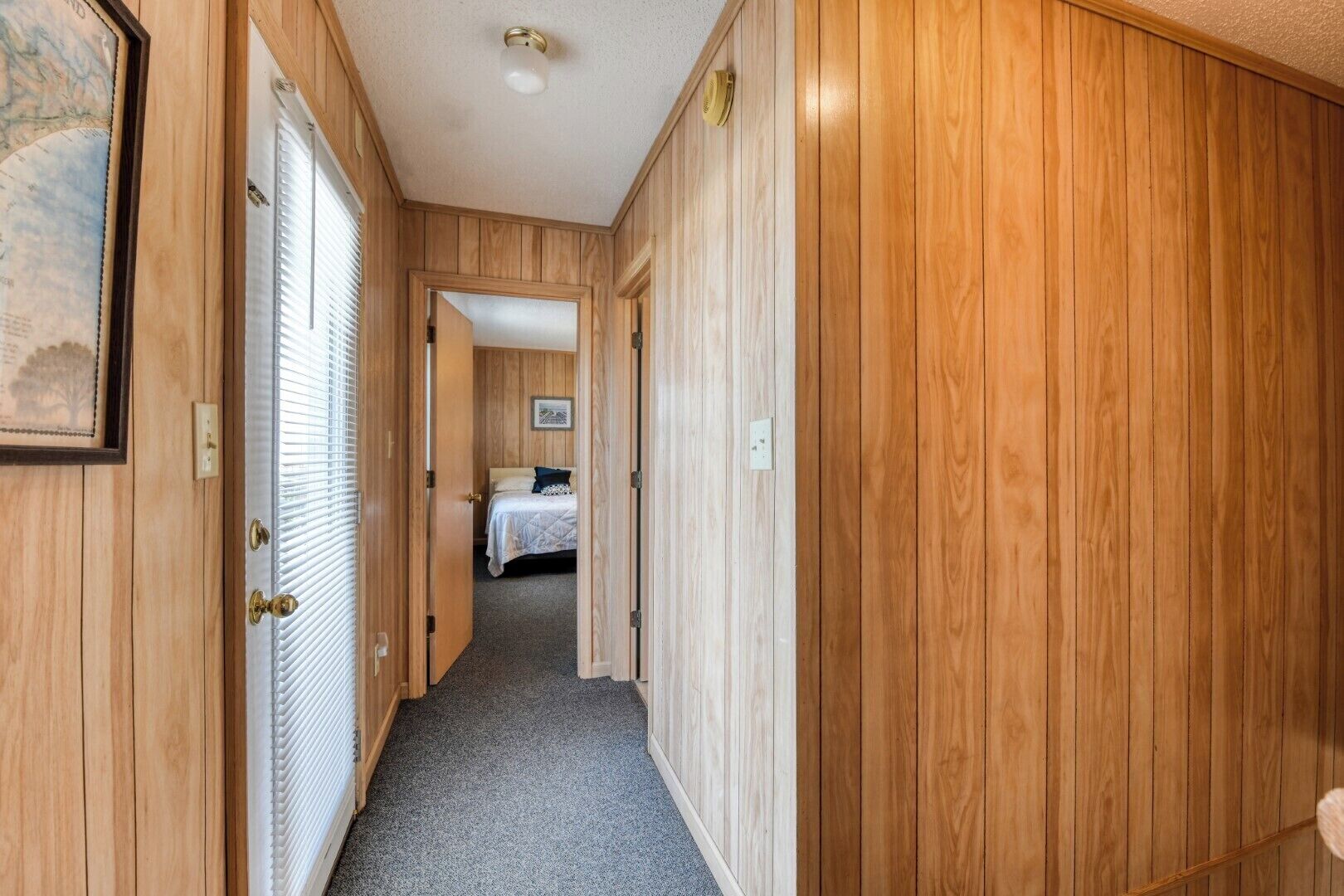 Un pasillo con paneles de madera, una cama con cubrecama blanco y una sola almohada, y un cuadro enmarcado en la pared izquierda.