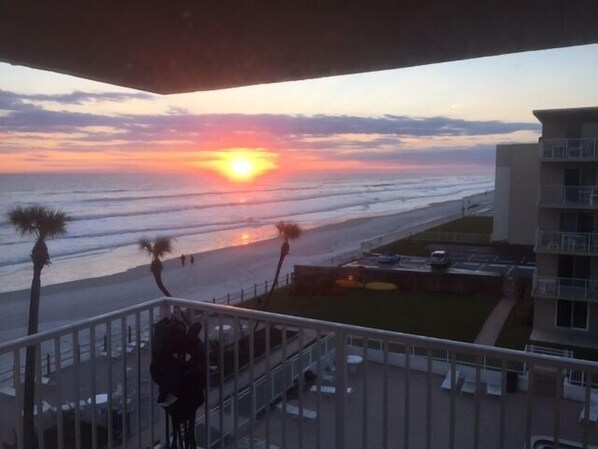Ein Balkon mit Blick auf den Sonnenuntergang über dem Meer, Palmen und einen Parkplatz.