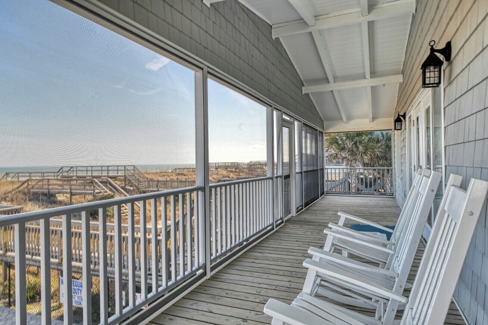 A covered balcony with white chairs, a view of the beach, and a railing.