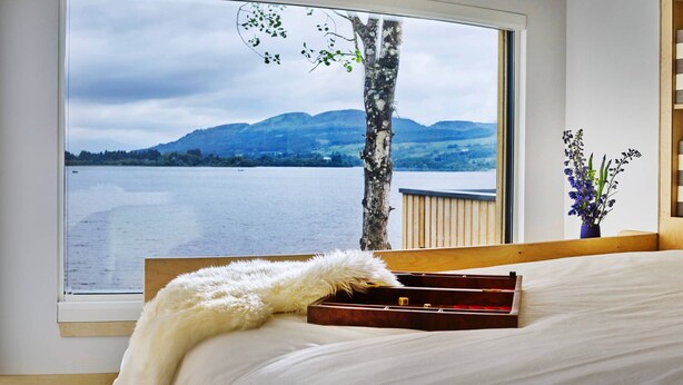 Une chambre avec une grande fenêtre donnant sur un lac et des montagnes. On y trouve un lit avec une literie blanche et un plaid blanc moelleux. Une table de chevet en bois, sur laquelle est posé un petit vase de fleurs, est visible.