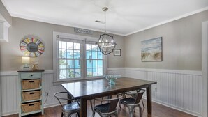 Spisestue med et træbord, fire stole, en lysekrone, et vindue med skodder, et skilt på væggen med teksten 'BEACH HOUSE' og et maleri.