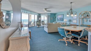 A spacious room with a large window overlooking the sea, a dining area with blue chairs, and a cozy seating area with a sofa and armchairs. - Surfmaster 103 - Three-bedroom, three-bath first-floor oceanfront condo (Garden City Beach)