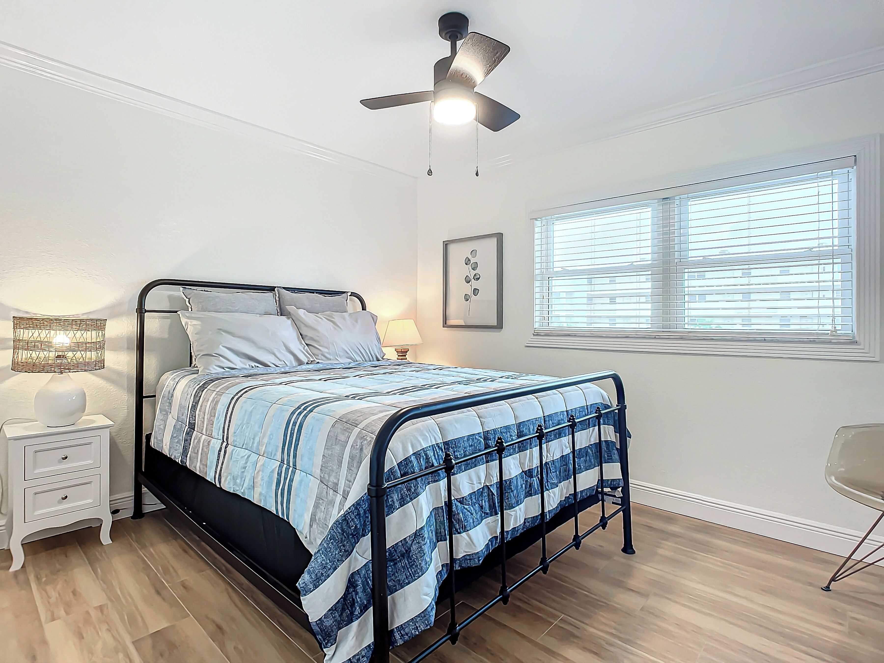 A bedroom with a bed, bedside tables, a ceiling fan, a window with blinds, and a framed picture on the wall.
