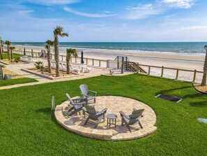 A patio area with Adirondack chairs and a fire pit, overlooking a beach with palm trees and a fence. - Watch the sunrise over the No Drive Beach in NSB! (New Smyrna Beach)