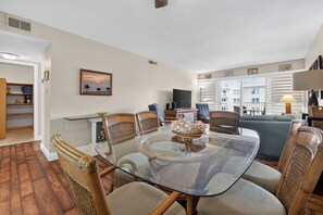 A dining area with a glass table surrounded by wicker chairs, a television, and a view of the outdoors through large windows.
