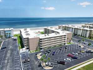 A multi-story hotel with a rooftop pool, parking lot, and a view of the beach.