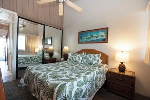 A hotel room with a bed, a nightstand, a lamp, a ceiling fan, a mirror, and a painting on the wall.