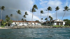 Un hôtel situé en bord de mer, avec un mur de pierre et des palmiers, un ciel dégagé et un arc-en-ciel bien visible.