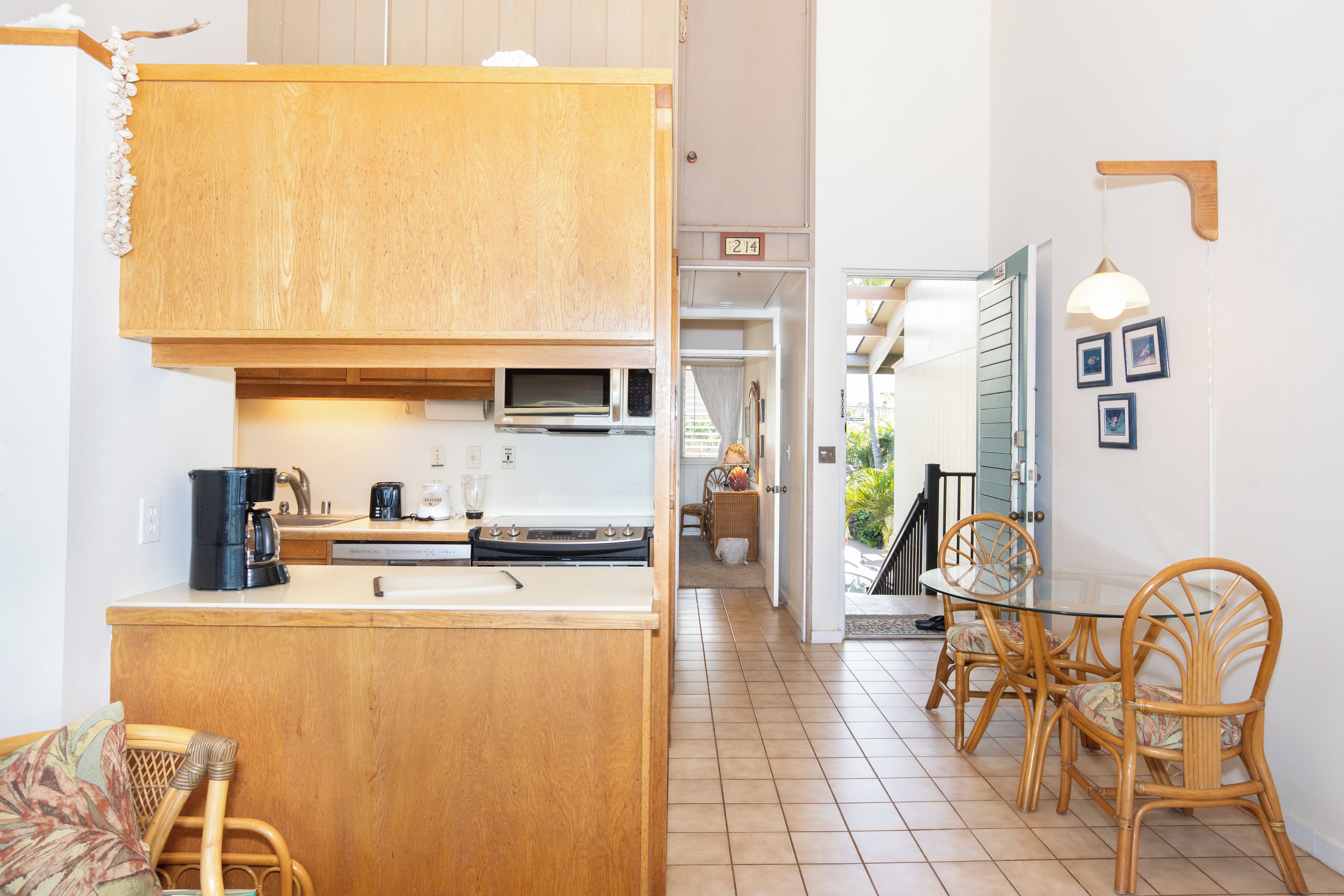 A compact kitchen with wooden cabinets, a microwave, and a coffee maker. There is a dining area with a glass table and wicker chairs.