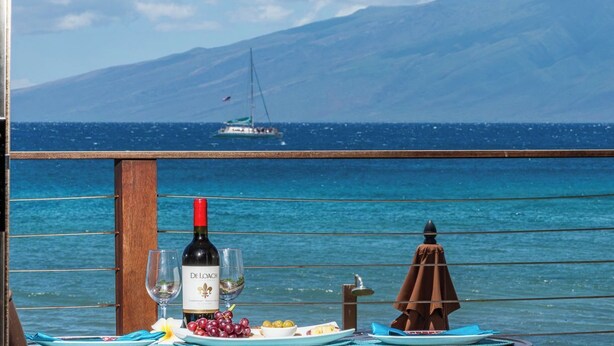 A table with a bottle of wine, a glass, and a plate of grapes, set against a scenic ocean view with a sailboat in the distance. - Top Floor! Direct Ocean View! Vaulted Ceilings! Winter Spec! #218 (Lahaina)
