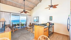 A spacious living area with a large sliding glass door leading to a balcony with a view of the ocean. There are wicker furniture pieces, a kitchen with a countertop, and ceiling fans. - Top Floor! Direct Ocean View! Vaulted Ceilings! Winter Spec! #218 (Lahaina)