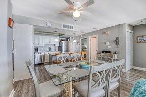 A modern kitchen with white cabinets, a dining area with a glass table and white chairs, and a ceiling fan.