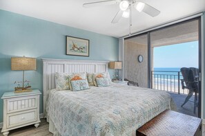 A bedroom with a bed, a nightstand, a lamp, a clock, and a view of the beach.