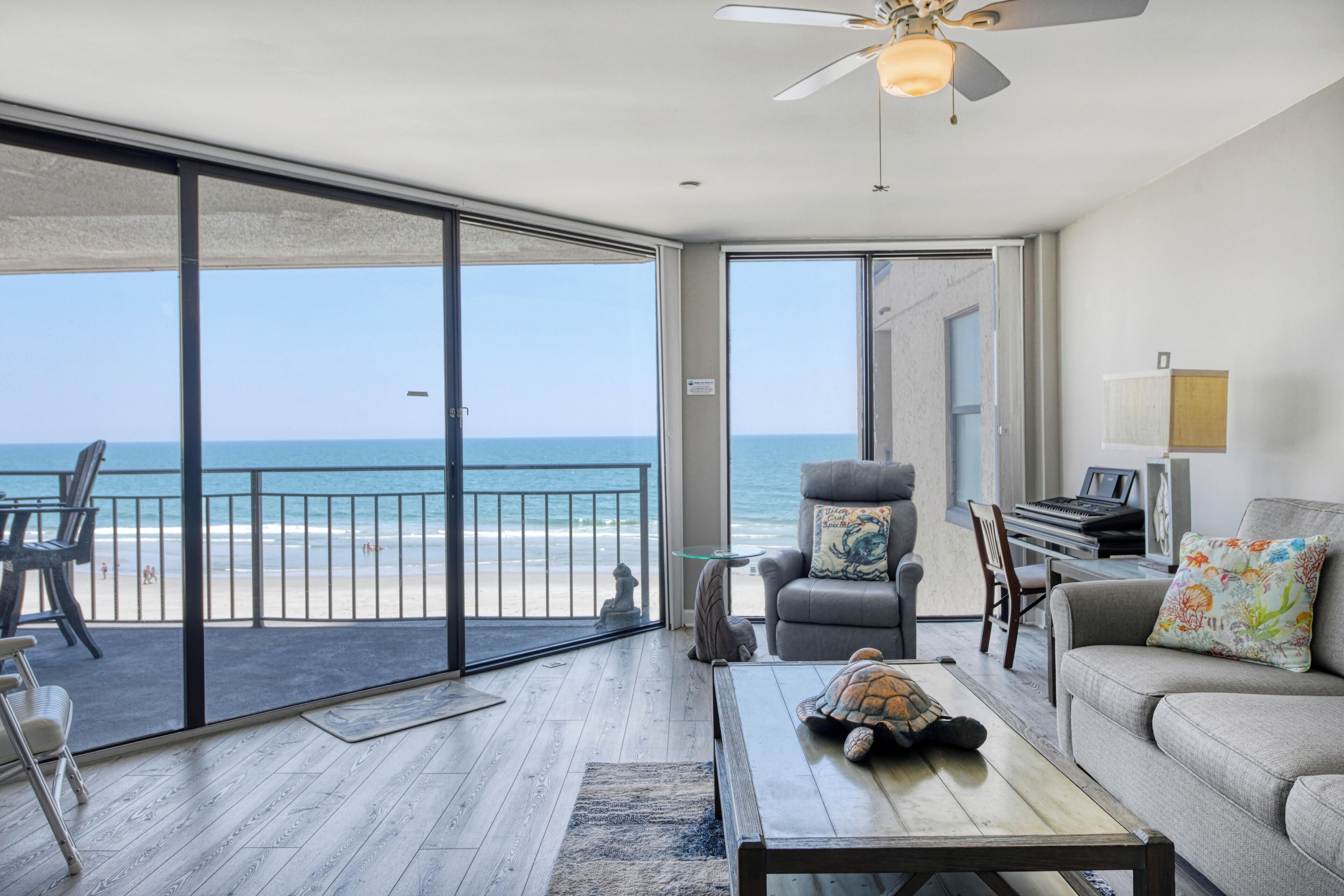 Une chambre avec vue sur la plage, un ventilateur de plafond, un piano et une tortue posée sur une table basse.